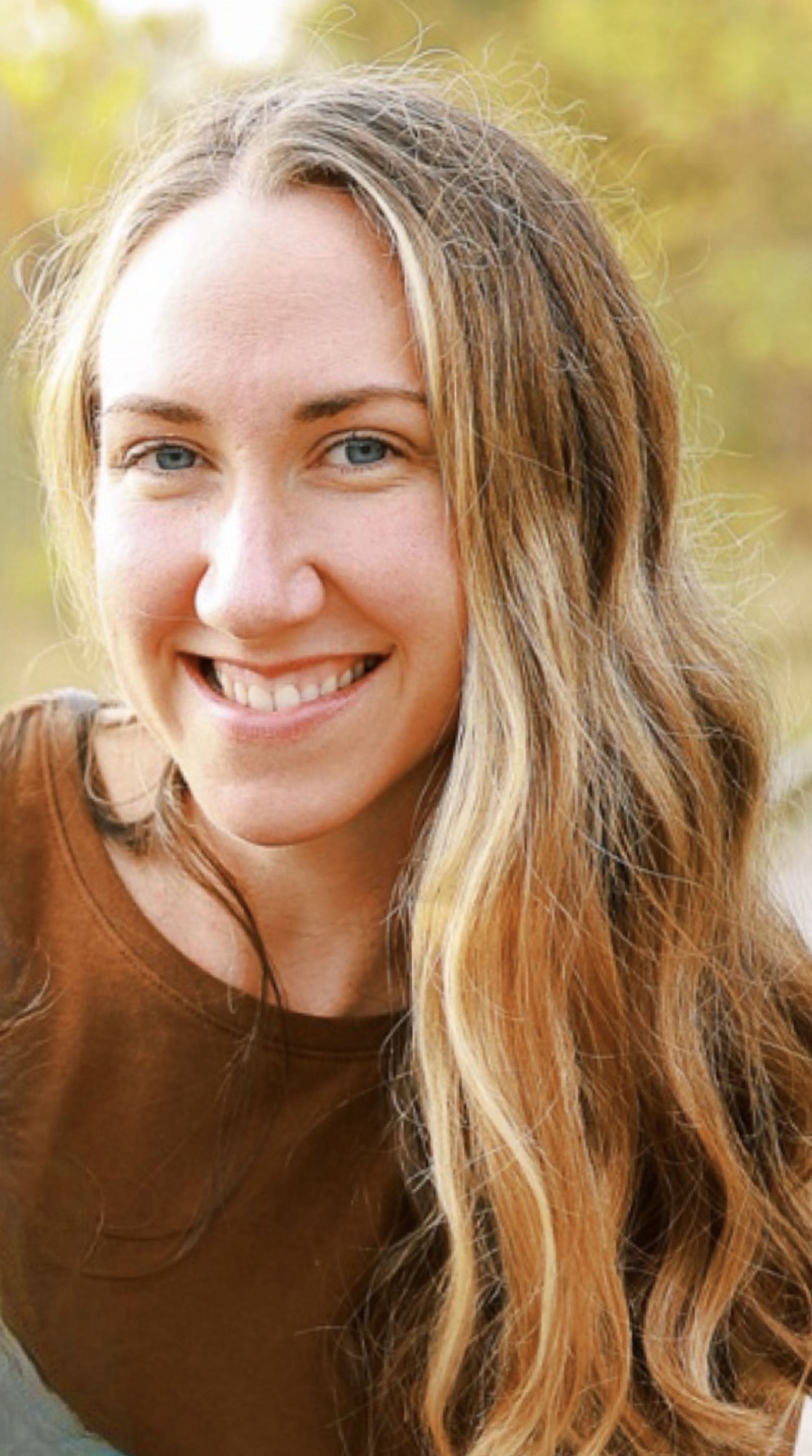 Close-up of a smiling person with long light brown and blonde hair, outdoors with a softly blurred green background
