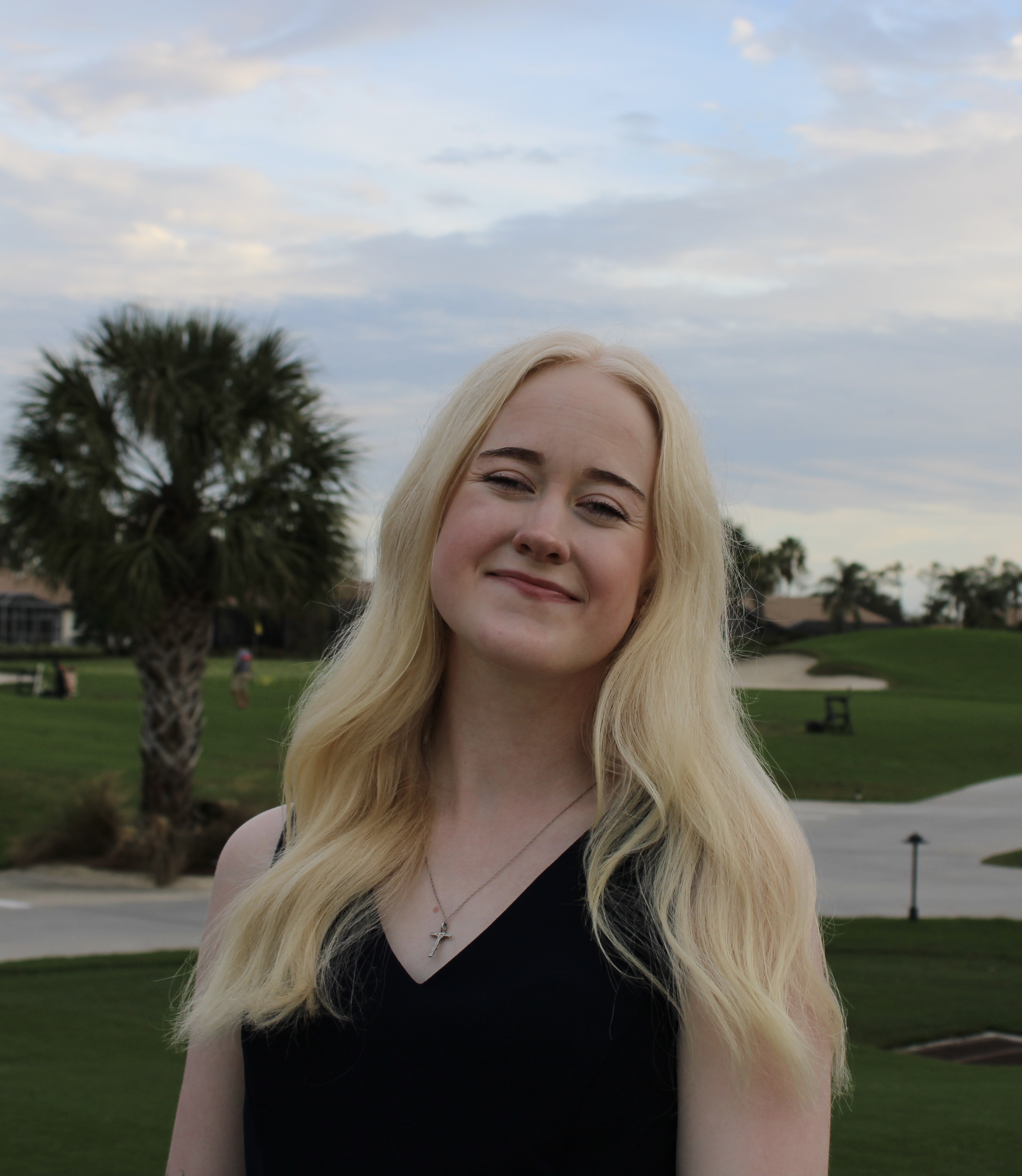 A person with long blonde hair smiling while standing outdoors on a grassy area with palm trees and a cloudy sky in the background